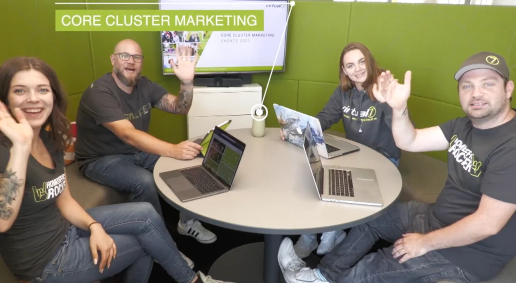 Vier Teammitglieder vom Core Cluster Marketing lächeln und winken in die Kamera, sitzen um einen Tisch mit Laptops im virtual7 Büro mit grüner Wanddekoration.