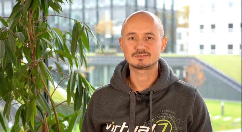 virtual7 Mitarbeiter Marcus steht im Büro neben einer grünen Pflanze, trägt einen grauen Hoodie mit virtual7-Logo und spricht lächelnd im Video über Responsiveness.